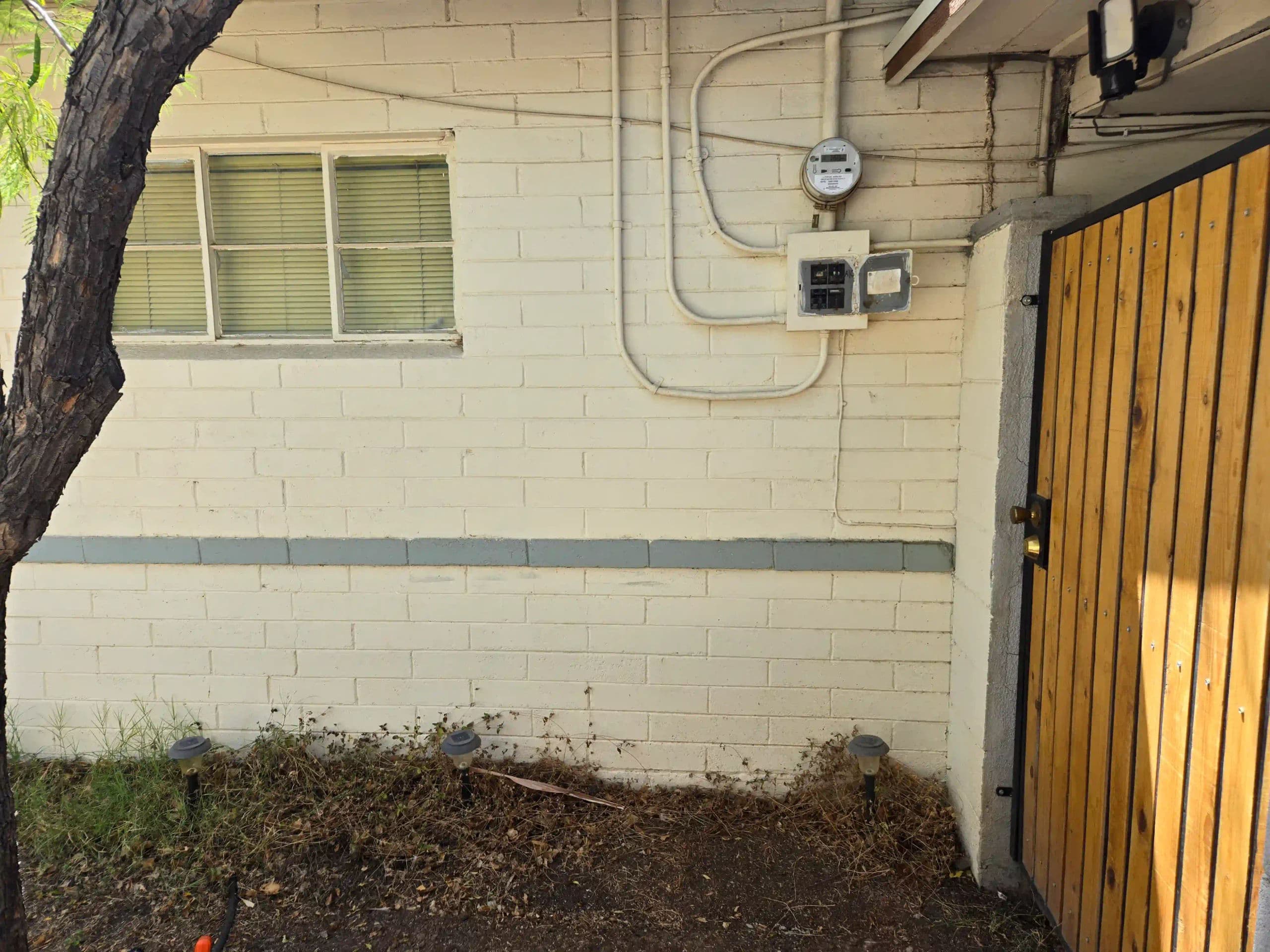 View of a building wall with a window and utility meters beside a wooden gate.