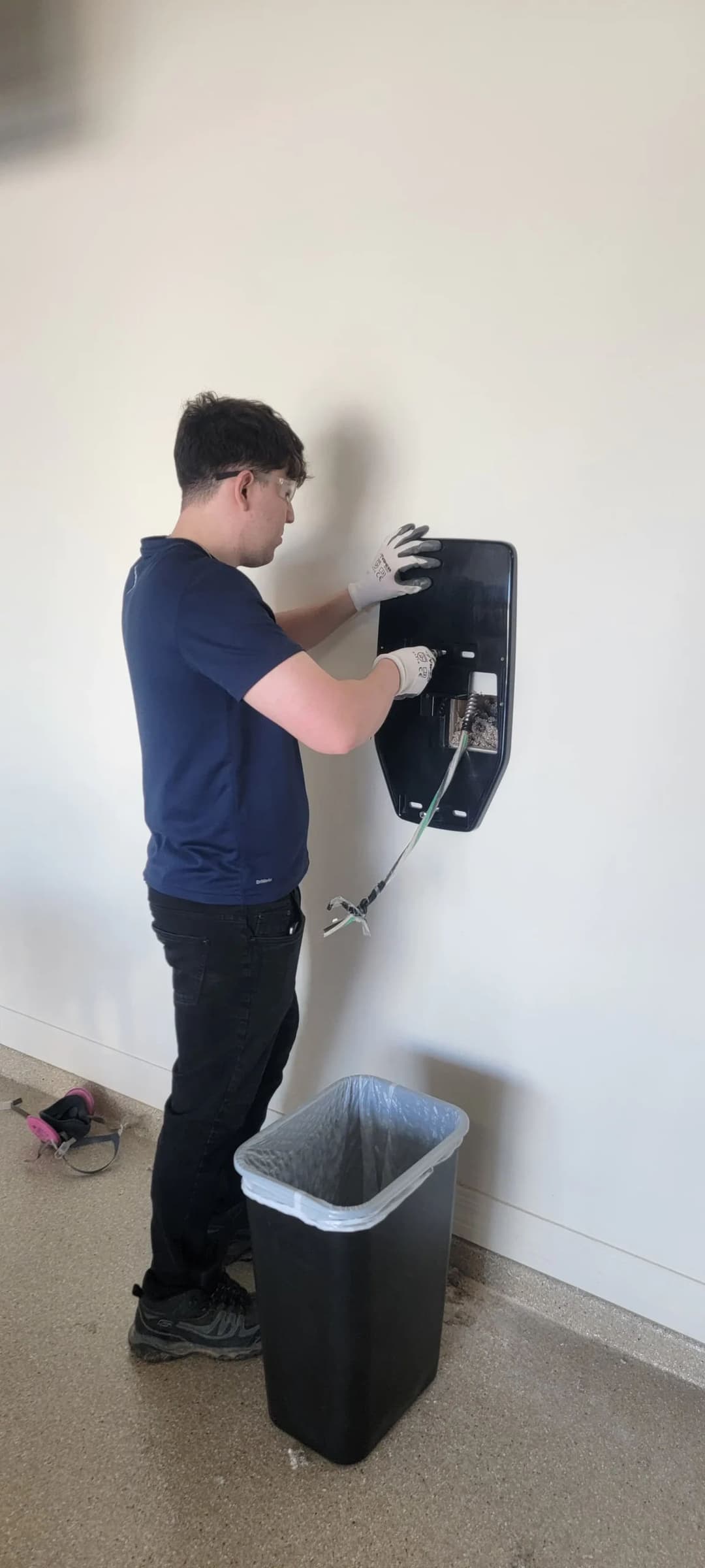 Person installing a wall-mounted device near a trash bin in a room setting.