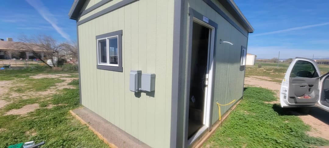 Exterior view of a small green shed with an open door and electrical connections.