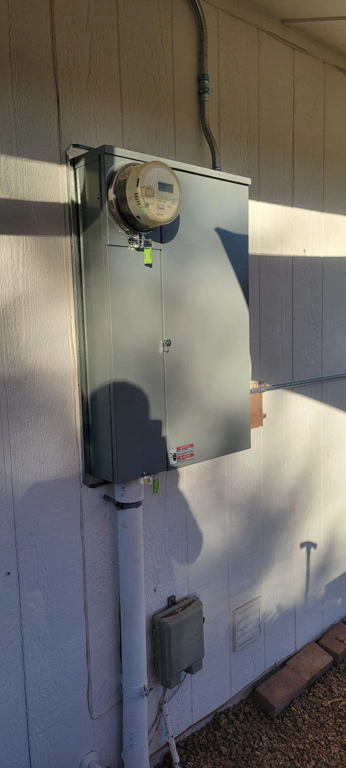 Electric meter mounted on exterior wall, with wiring and utility box visible underneath.