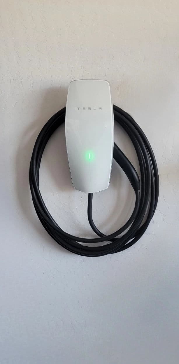 Tesla wall-mounted EV charger with green indicator light and charging cable.