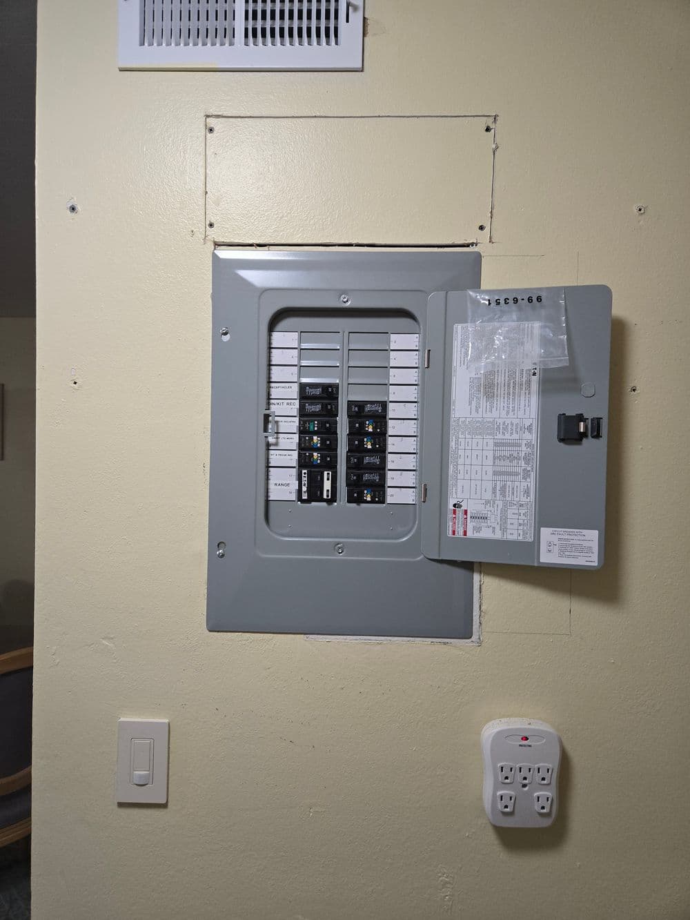 Electrical panel on a beige wall with exposed area, nearby power outlet visible.