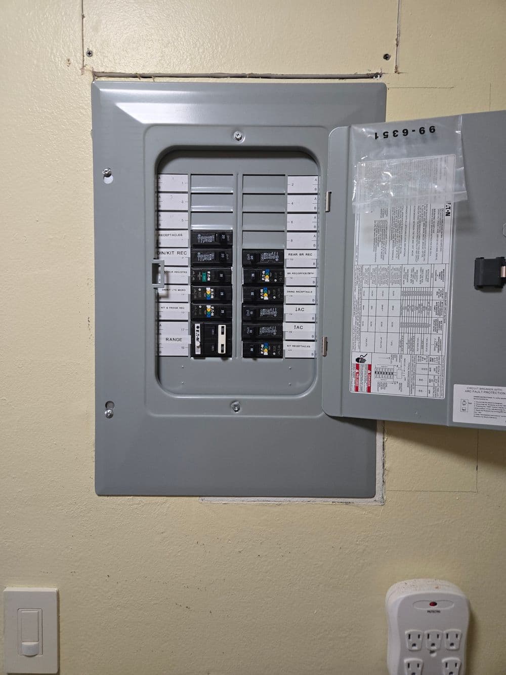 Electrical panel with circuit breakers, mounted on a beige wall, showing various labels and switches.