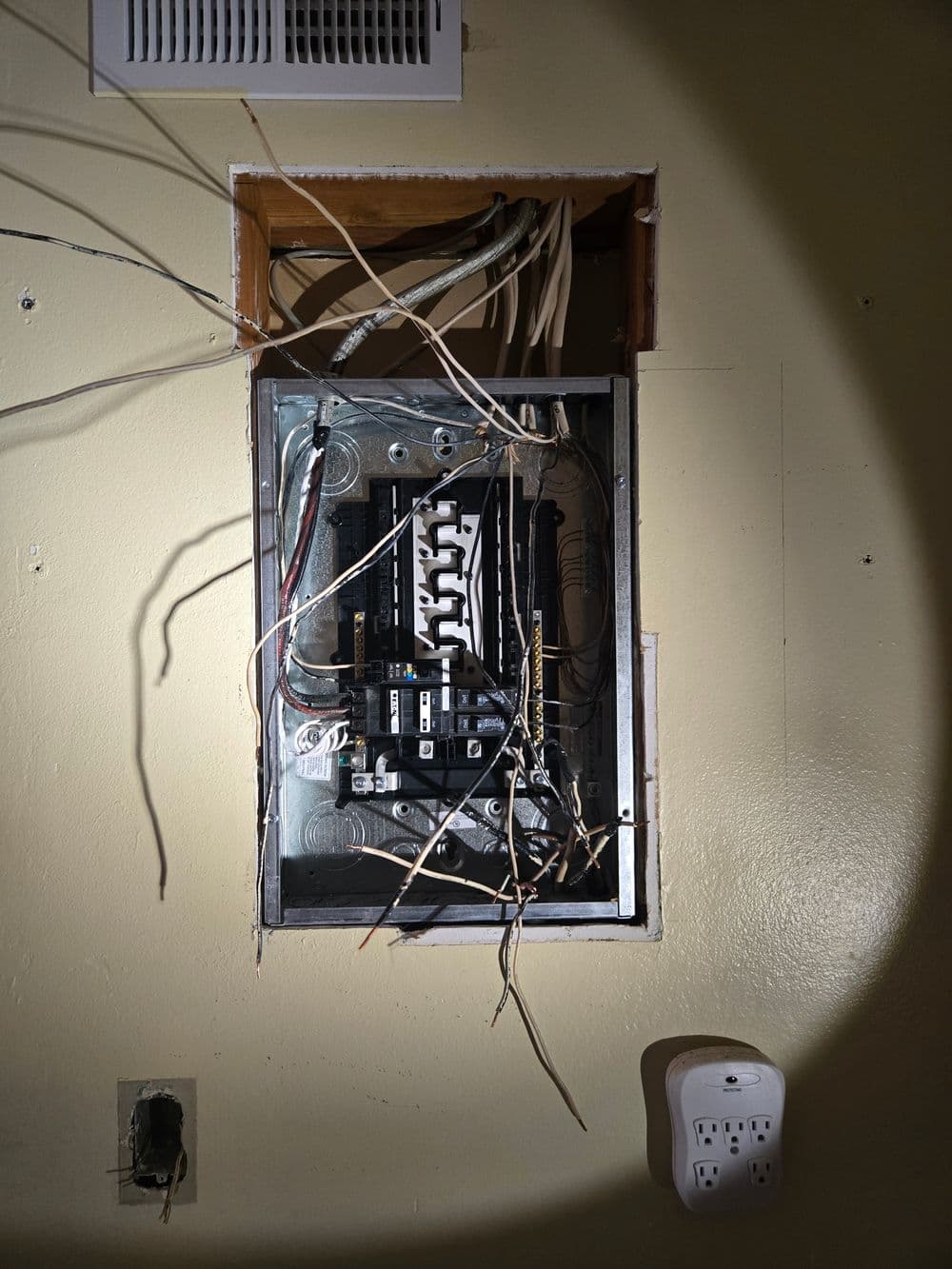 Electrical panel with exposed wires, inside a wall, illuminated by a flashlight.