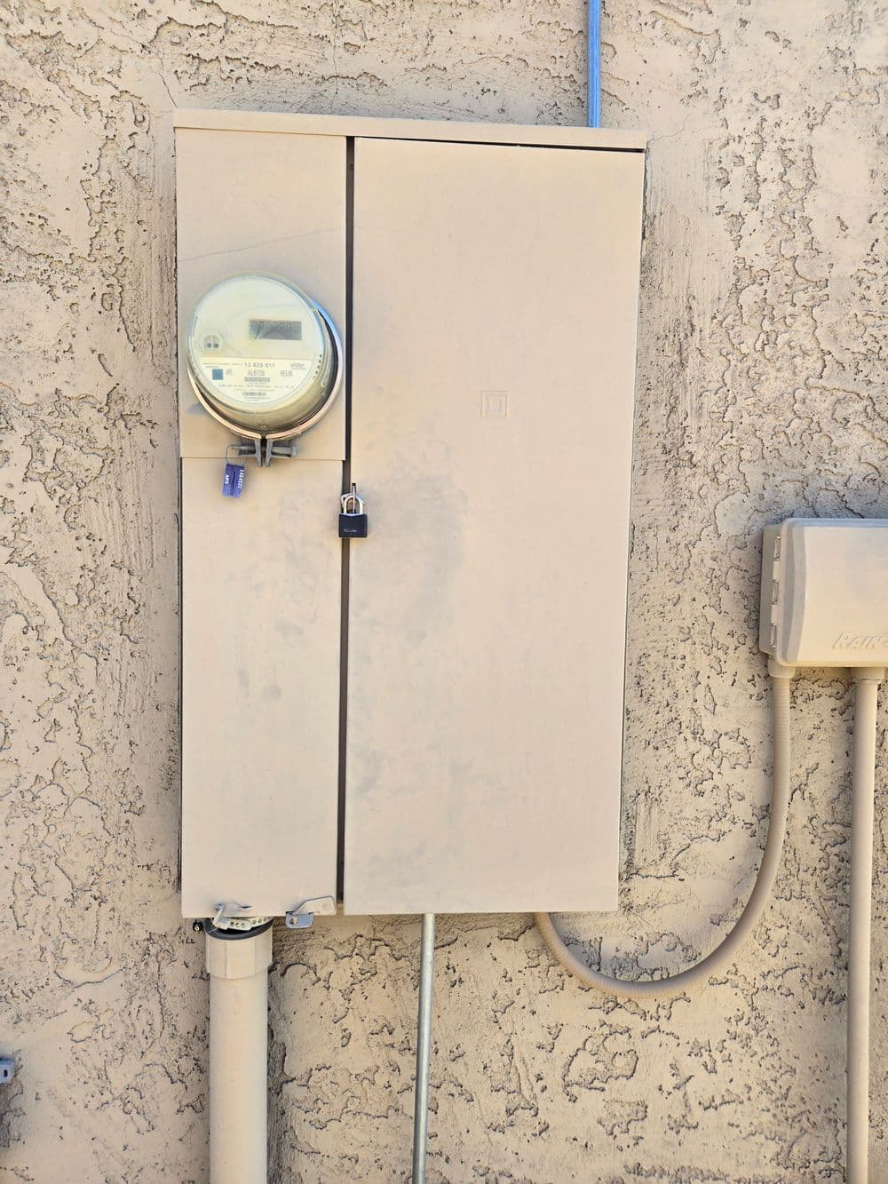 Electric meter mounted on a beige wall with a locked cabinet and utility connections.