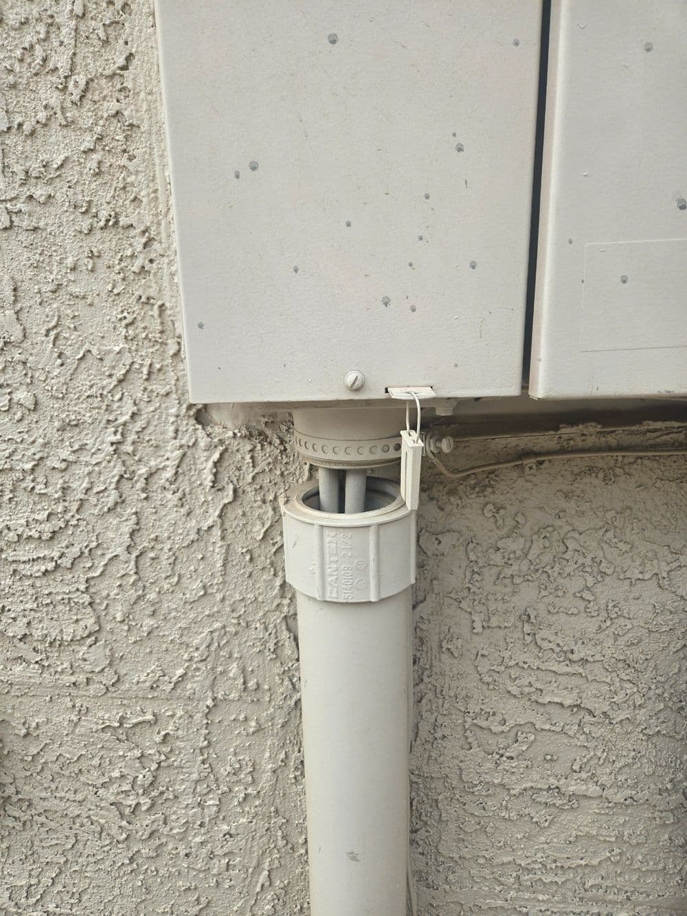 Gray utility box mounted on textured wall with a white drainage pipe below.