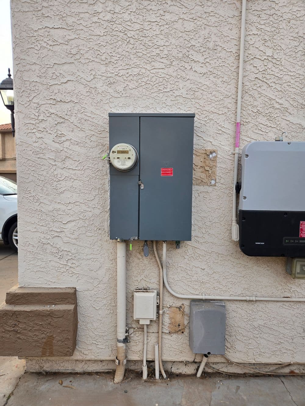 Electric meter box on a residential wall with utility connections and a nearby inverter.