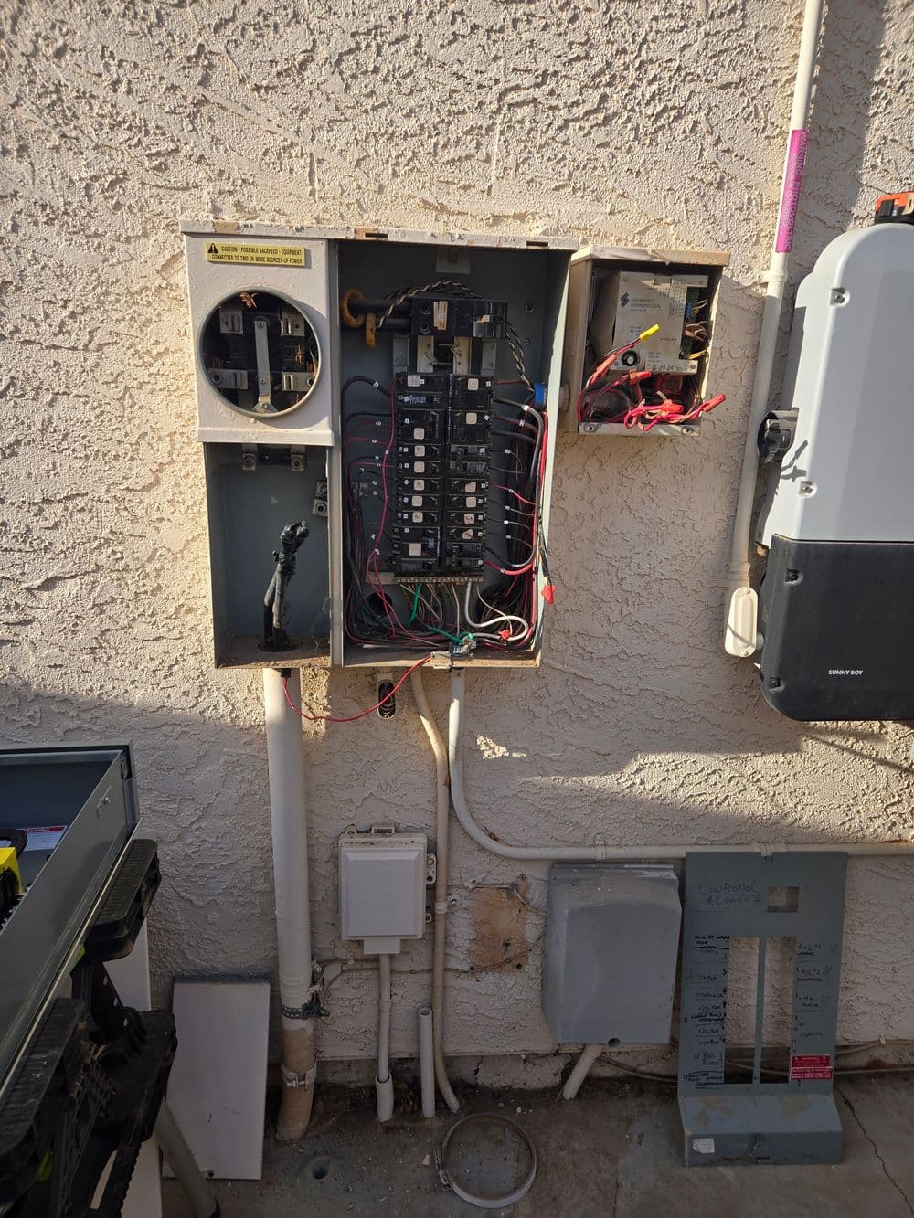 Electrical panel with exposed wiring and circuit breakers mounted on a textured wall.