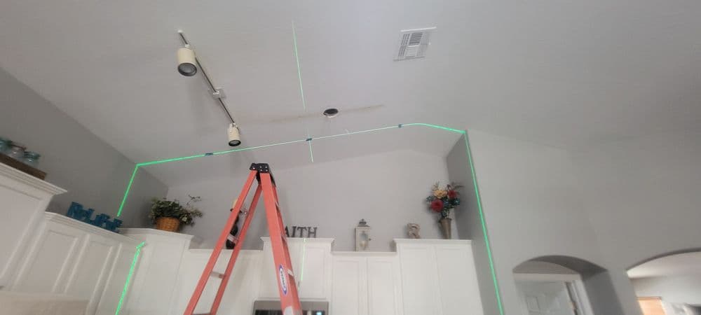 Laser level tool projected on kitchen ceiling above a ladder, with cabinets and decorative items.
